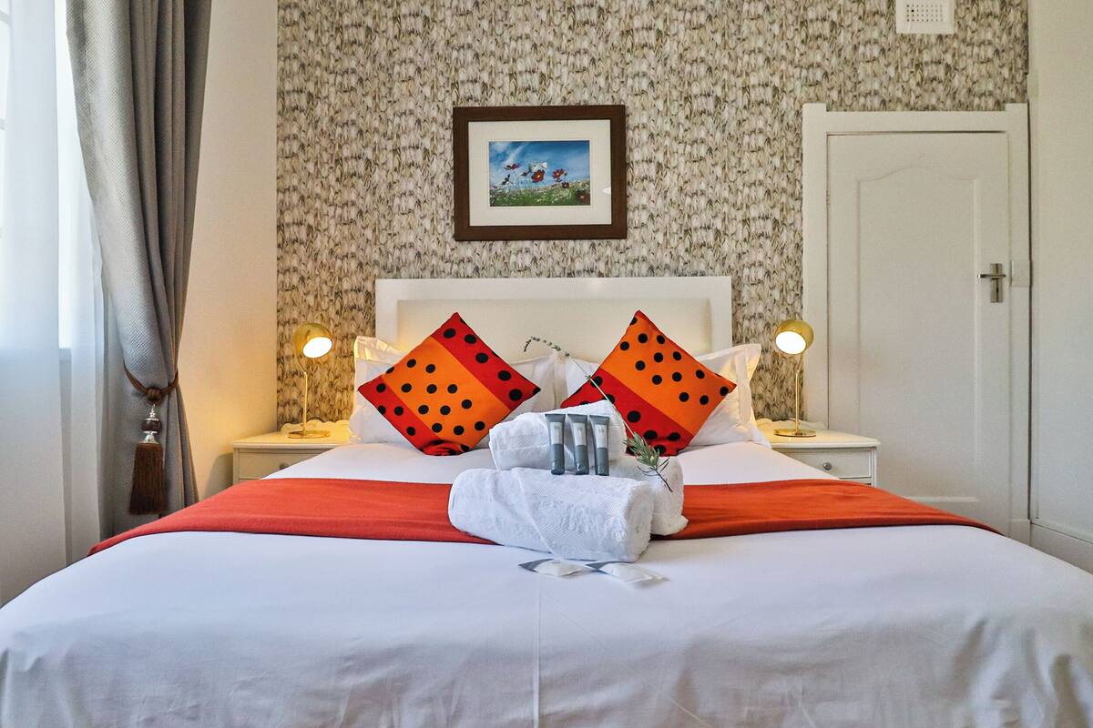 Ein ordentlich bezogenes Bett mit einer weiß-roten Tagesdecke, zwei Kissen mit schwarzen Punkten und ein Handtuch auf dem Bett. Zwei Nachttische mit Lampen, ein gerahmtes Bild an der Wand und eine Tür auf der rechten Seite sind ebenfalls zu sehen.