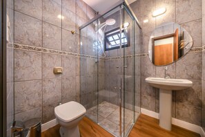 A bathroom with a glass-enclosed shower, a toilet, and a sink with a mirror. - Casa alto padrão com piscina 15 pessoas (Bombinhas)