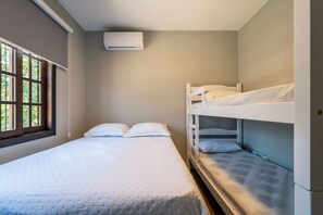 Una cama individual con cubrecama acolchado, una ventana con persianas y una litera con ropa blanca de cama.