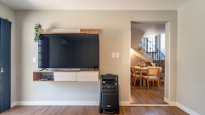 Una stanza con una TV a schermo piatto montata a parete, un tavolo da pranzo in legno con sedie e una valigia sul pavimento.