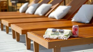 A row of wooden lounge chairs with white cushions, a can of drink, and sunglasses on a wooden table.