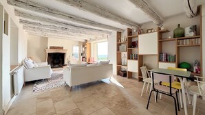 Ein gemütliches Wohnzimmer mit Kamin, einem weißen Sofa, einem Esstisch mit Stühlen und Holzböschern.