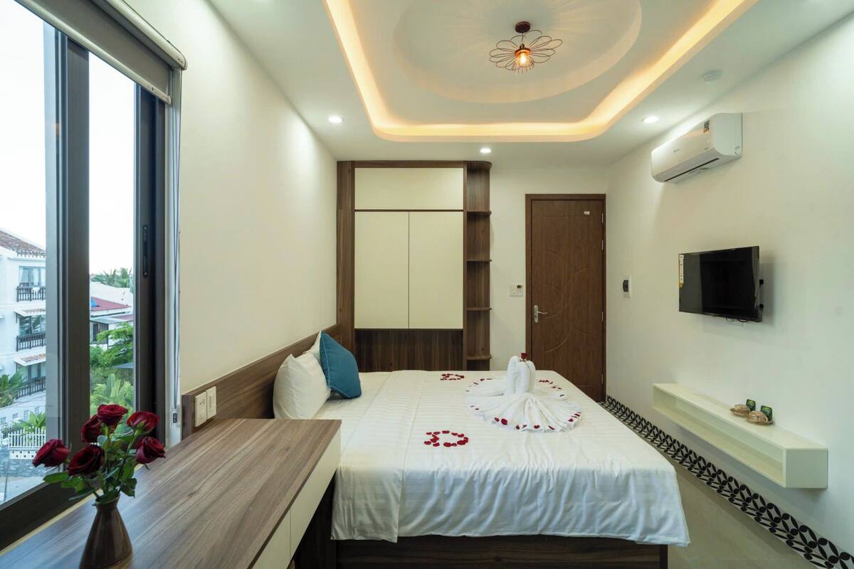 A hotel room with a bed, a TV, a window, and a bedside table with a vase of flowers.