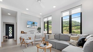 A spacious living room with a grey sofa, wooden chairs, a coffee table, and large windows offering a view of greenery. - Sophisticated Modern Home w/ Ocean Views, Gorgeous Master Suites & Private Jacuzzi (Las Catalinas)