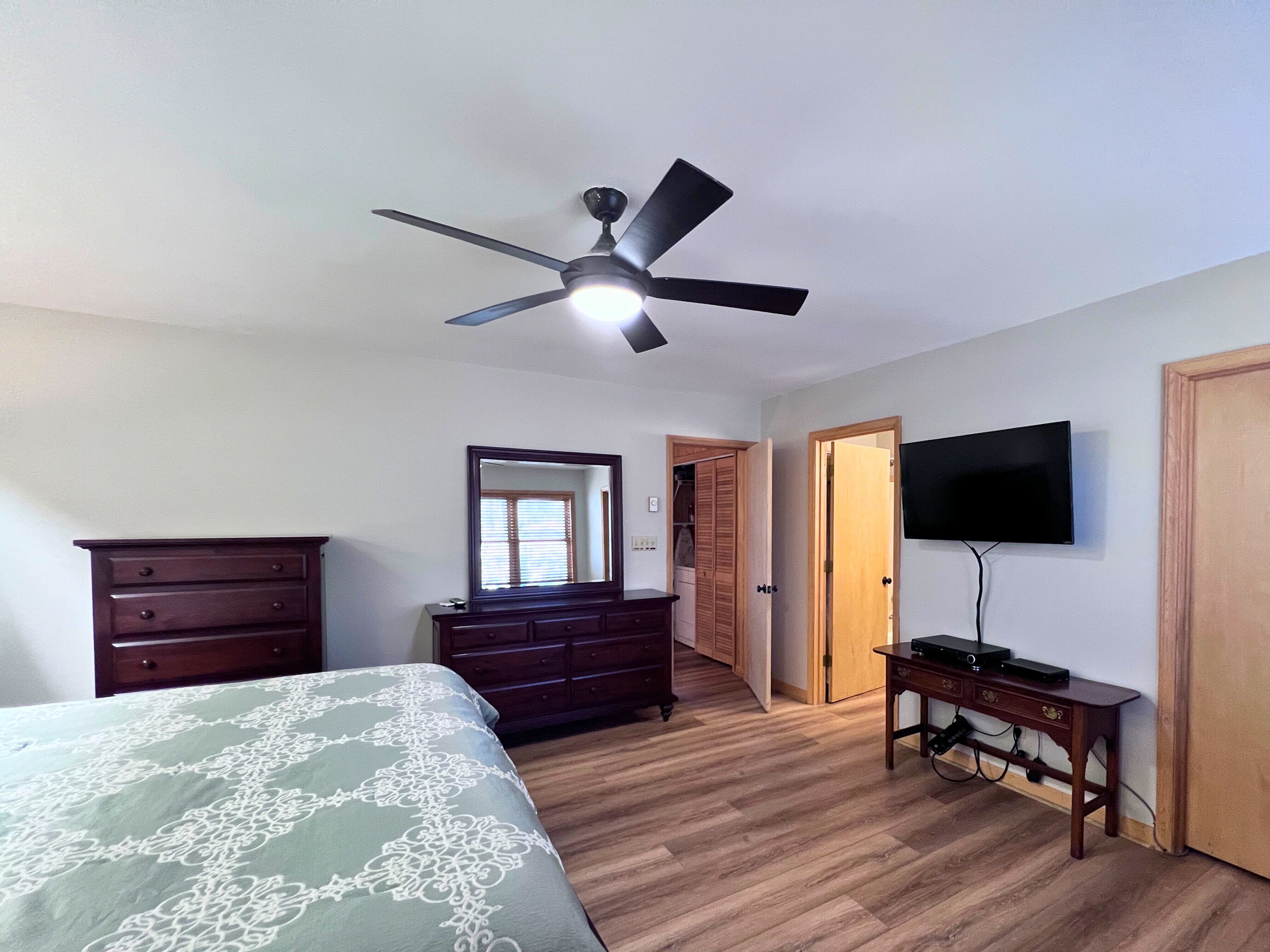 A bedroom with a bed, dresser, mirror, ceiling fan, TV, and wooden flooring.