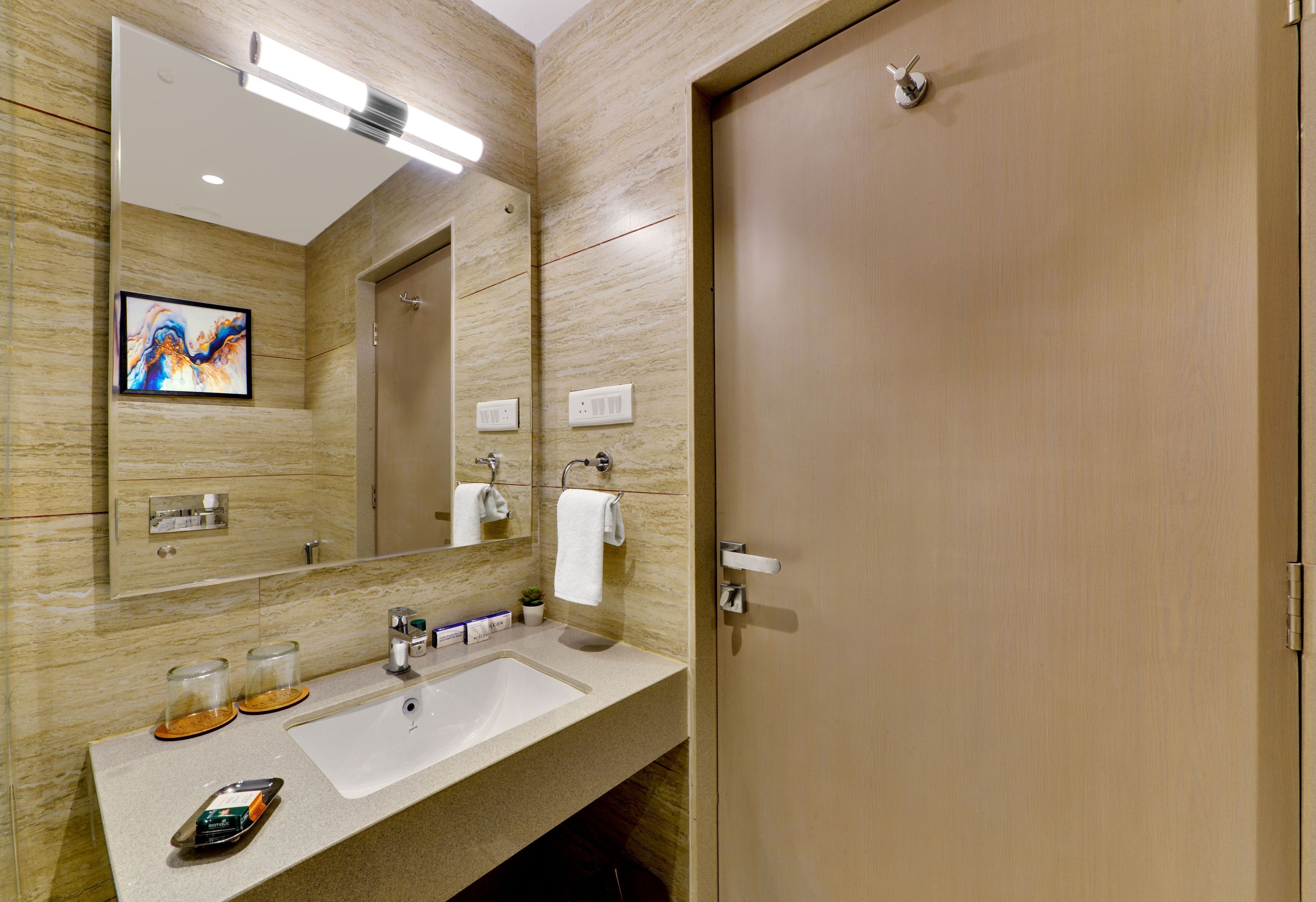 Une salle de bain moderne avec un mur en marbre beige, un grand miroir, un lavabo doté d’un distributeur de savon en verre et une porte en bois.