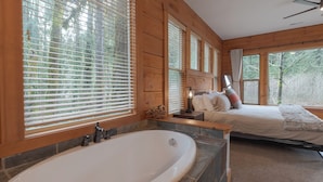 Une chambre située dans un chalet en bois, avec une grande baignoire, un lit avec des oreillers et une vue sur les arbres à travers de grandes fenêtres.