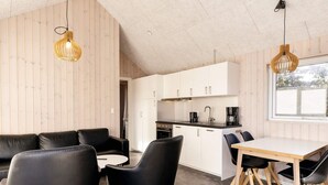 A modern kitchen with white cabinets, a black countertop, and a coffee maker. There is a dining area with a wooden table and black chairs. - 5 Star Holiday Home in Lokken (Lokken)