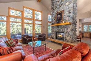 A spacious living room with leather sofas, a stone fireplace, large windows, and a view of trees.