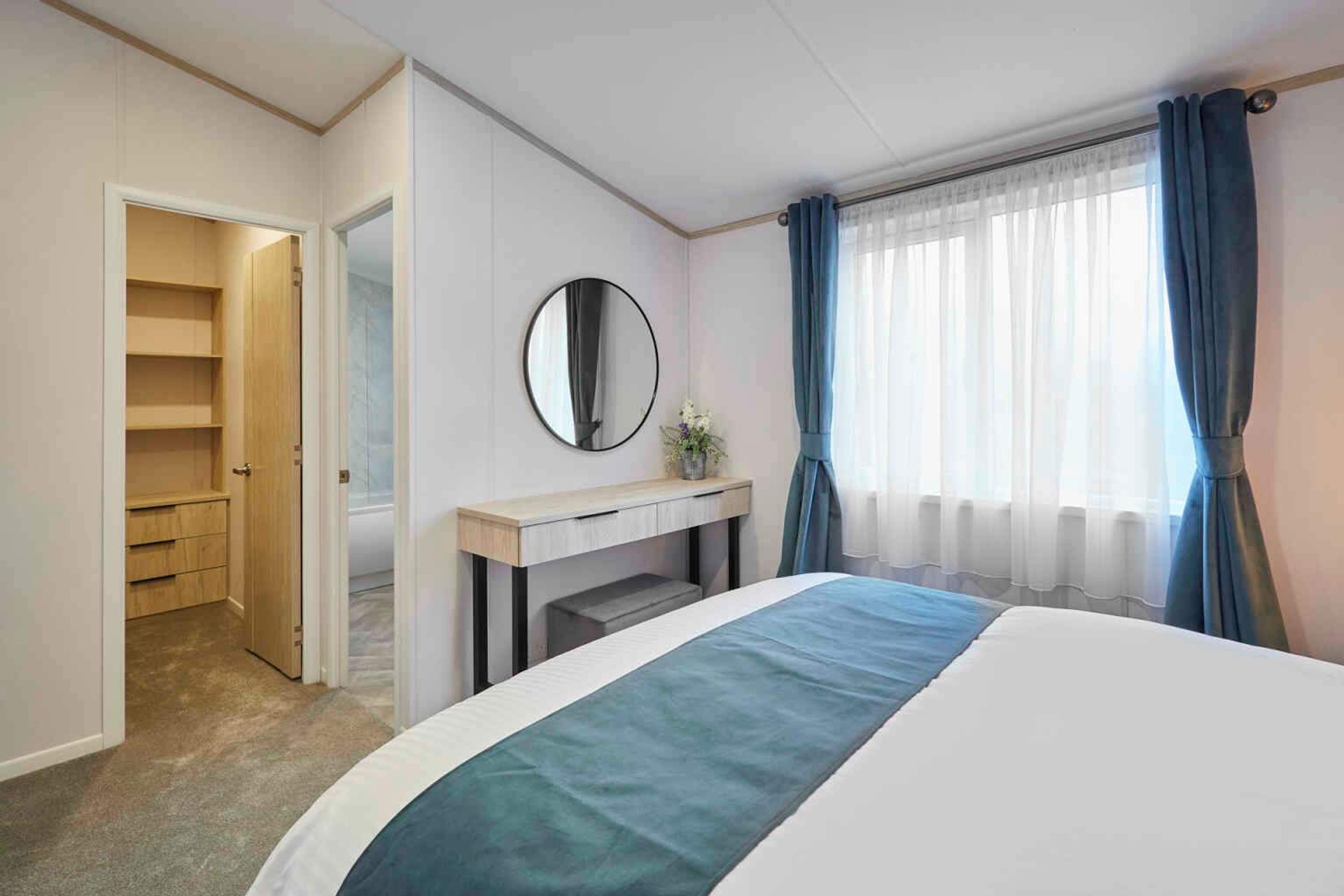 A hotel room with a bed, a bedside table with a mirror, a window with curtains, and a walk-in closet.