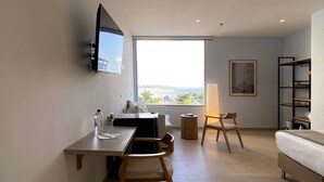 Et moderne hotelværelse med en seng, et skrivebord med stol, et fjernsyn ophængt på væggen, et stort vindue med udsigt og en reol.