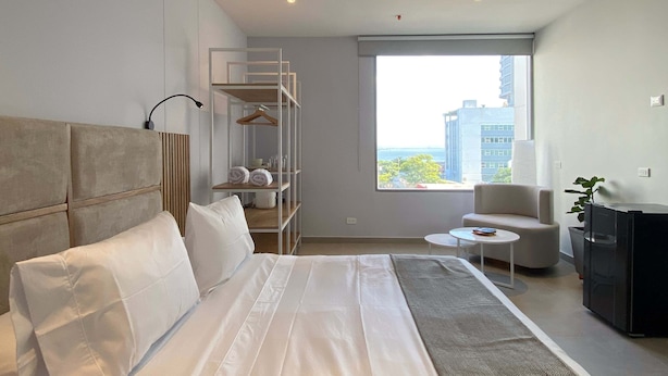 Et moderne hotelværelse med en stor seng, et siddeområde med en stol og et lille bord, en indbygget garderobe og et stort vindue med udsigt over byen.