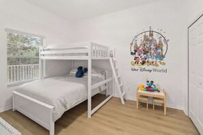 A child's room with bunk beds, a Disney-themed wall decal, a small table with toys, and a window with blinds.