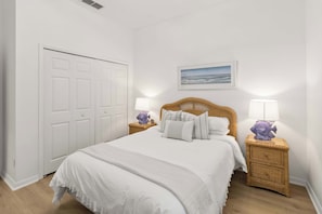 Un dormitorio ordenado con una cama, dos mesitas de noche, un cabecero de mimbre, una puerta blanca y un cuadro en la pared.