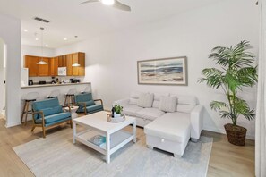 Una sala de estar moderna con un sofá blanco, sillas azules, una mesa de centro y una zona de cocina con armarios de madera y un microondas.
