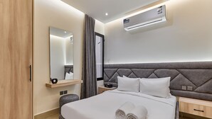 A modern hotel room with a large bed, a bedside table, a nightstand, a mirror, an air conditioning unit, and a wardrobe. - bnbme | Riviera-A5 | 3 Bdr (Riyadh)