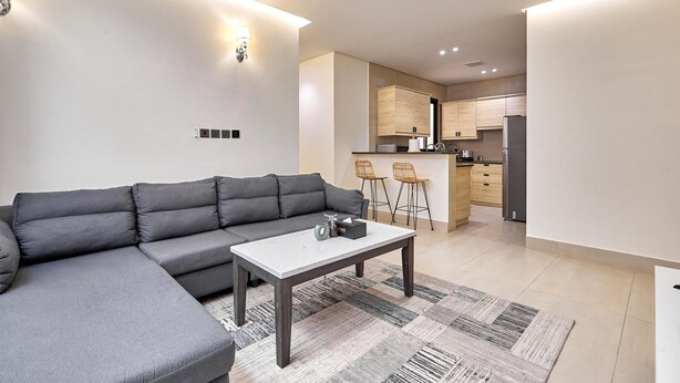 A modern living room with a grey sectional sofa, a white coffee table, and a kitchen area with wooden cabinets and a refrigerator. - bnbme | Riviera-A5 | 3 Bdr (Riyadh)