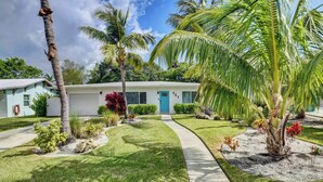 A single-story house with a white exterior, a blue door, and a well-maintained lawn with palm trees and shrubs. - Coastal Cottage private oasis w/ Hot Tub & less than 5 minutes to beach! (Boynton Beach)