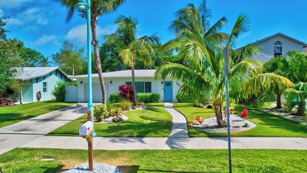A two-story building with a light blue exterior, a front porch, and a well-maintained lawn with palm trees and shrubs. - Coastal Cottage private oasis w/ Hot Tub & less than 5 minutes to beach! (Boynton Beach)