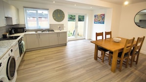 A modern kitchen with a built-in washing machine, a dining area with a wooden table and chairs, and a large wall clock. - 3 Bed, Sleeps 5, Pet Friendly, Free Parking (Mexborough)