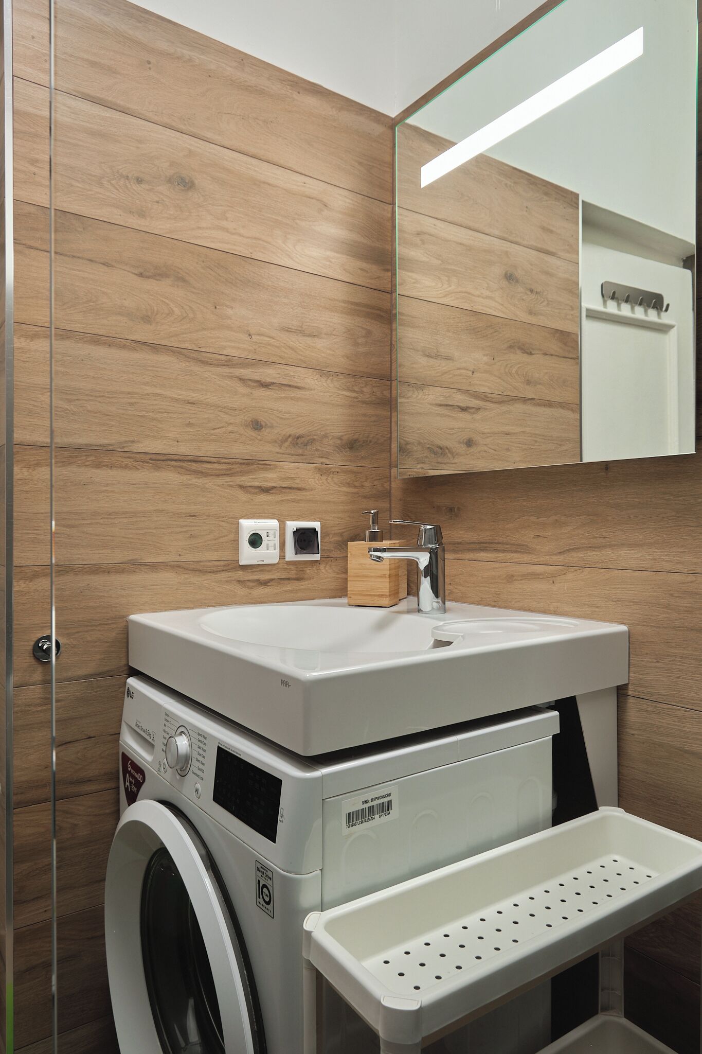 Ein modernes Badezimmer mit einer Holzwand, einem weißen Waschbecken, einer Waschmaschine und einem Spiegel.