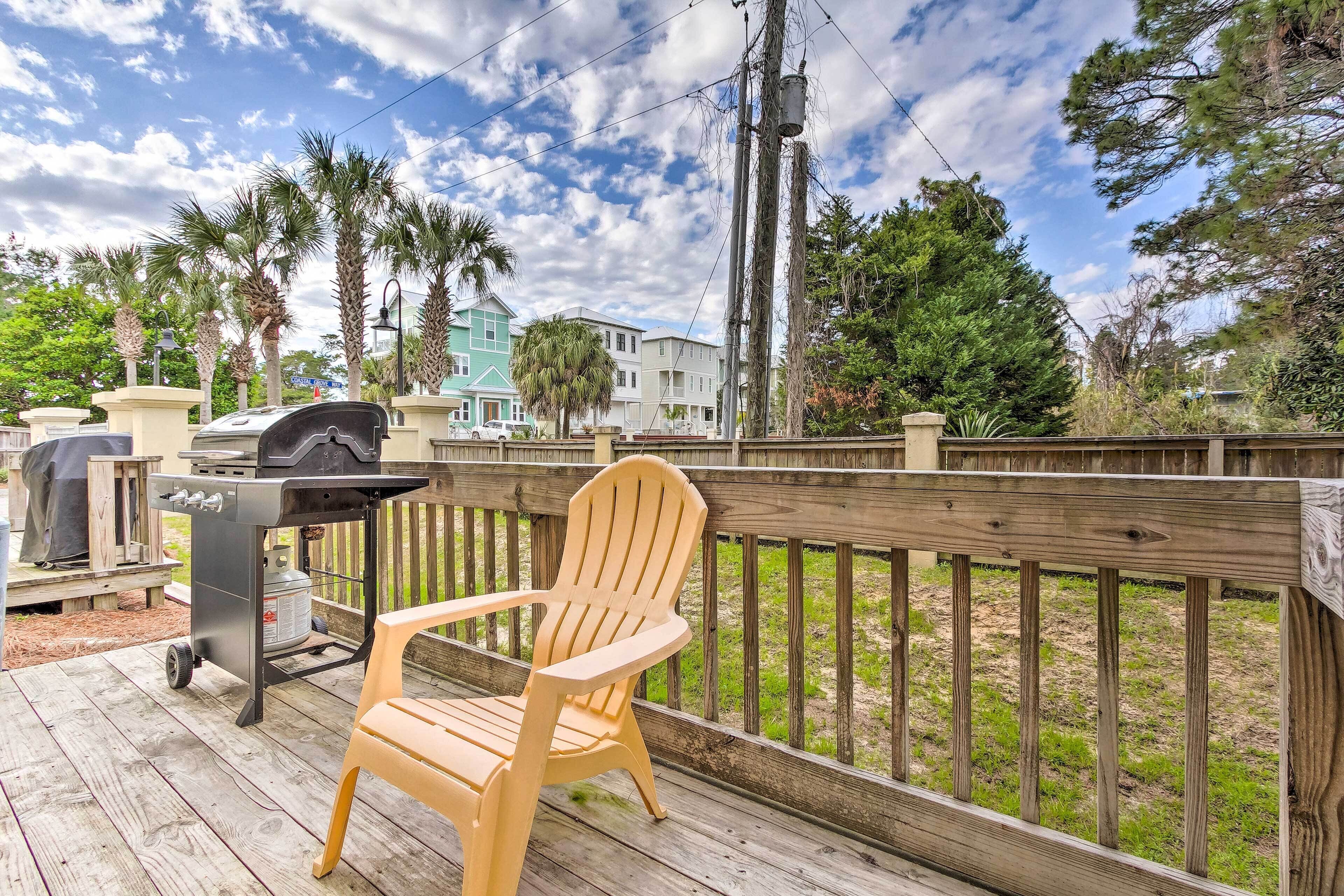 En træterrasse med grill og en stol, med udsigt over en græsplæne med træer og huse.