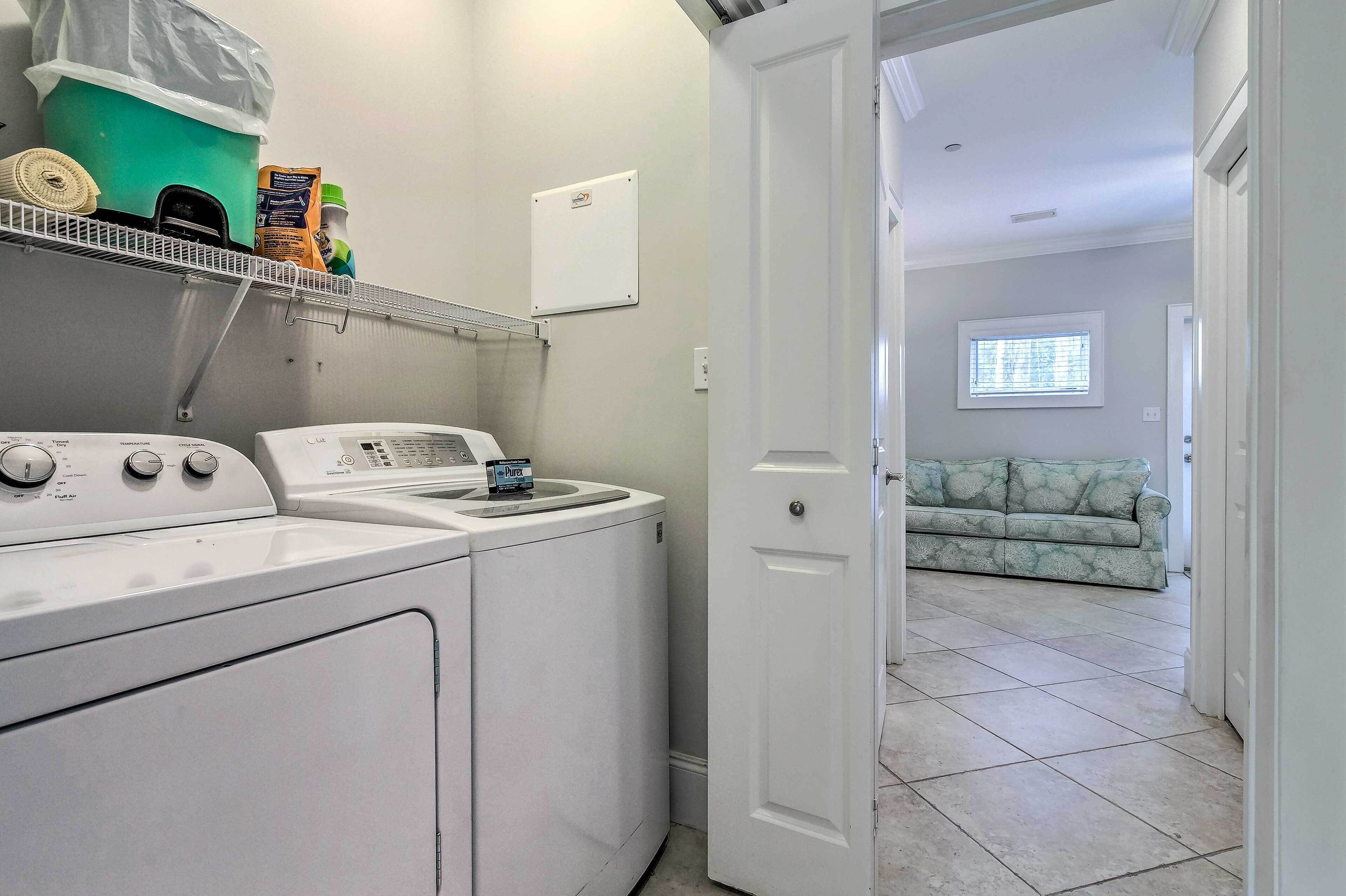 Un área de lavandería con lavadora, secadora y un estante con productos de limpieza. Una puerta da acceso a una sala de estar con sofá y ventana.