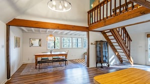 A spacious room with a wooden staircase, a dining area with a table and chairs, and large windows allowing natural light.