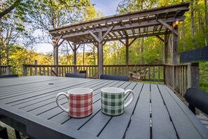 Una terraza de madera con una pérgola, dos tazas a cuadros sobre una mesa, y un fondo boscoso.