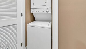 Una lavadora compacta y blanca en un lavadero pequeño con una pared beige y una puerta blanca con persianas horizontales.