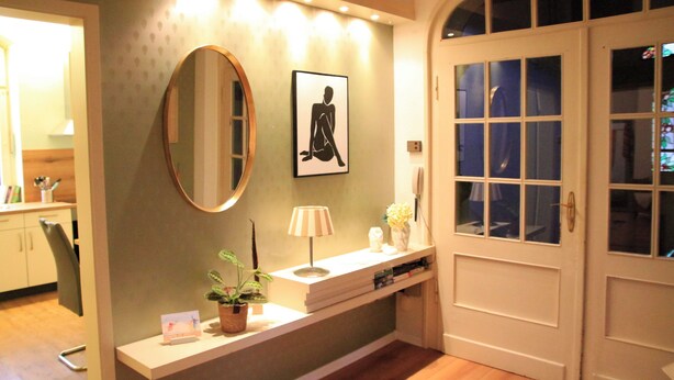 Un couloir bien éclairé, avec un miroir, un tableau encadré, une petite table avec une lampe et une plante en pot.