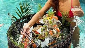 Iemand in een rood badpak die een drankje vasthoudt en een mand met sushi bij een zwembad.