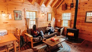 A cozy cabin interior with wooden walls, a sofa, a table, and a fireplace. - Cabin, Queen Bed, 2 Bunks, Private Bath (Greenville)