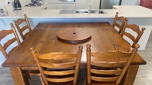 A wooden dining table with six chairs, a central decorative piece, and a kitchen area in the background.