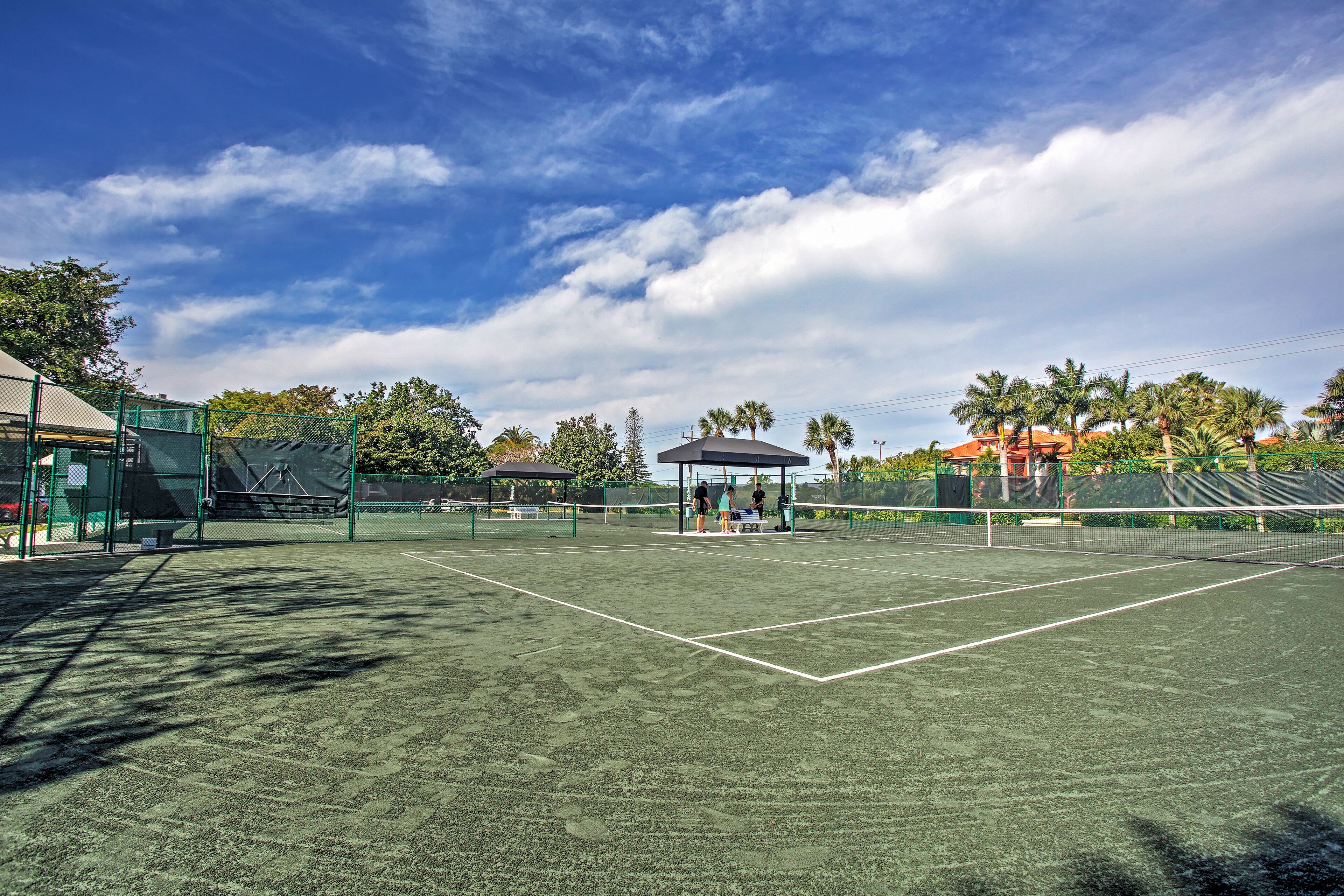 A tennis court with a pavilion, surrounded by trees and a fence.