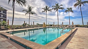 A swimming pool with a tiled edge, surrounded by lounge chairs and palm trees, with a building in the background.