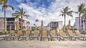 A poolside area with lounge chairs and umbrellas, surrounded by palm trees and modern buildings.