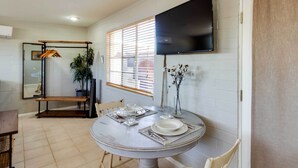 Un coin repas avec une table ronde prévue pour quatre personnes, un téléviseur fixé au mur et une fenêtre équipée de stores.