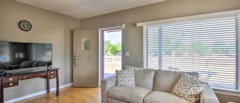 A living room with a flat-screen TV on a wooden desk, a beige sofa with patterned cushions, and a large window with blinds.