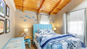 Une chambre avec un plafond en bois, un lit avec une literie bleue, une table de chevet avec une lampe et une petite table sur laquelle est posé un cadre photo.