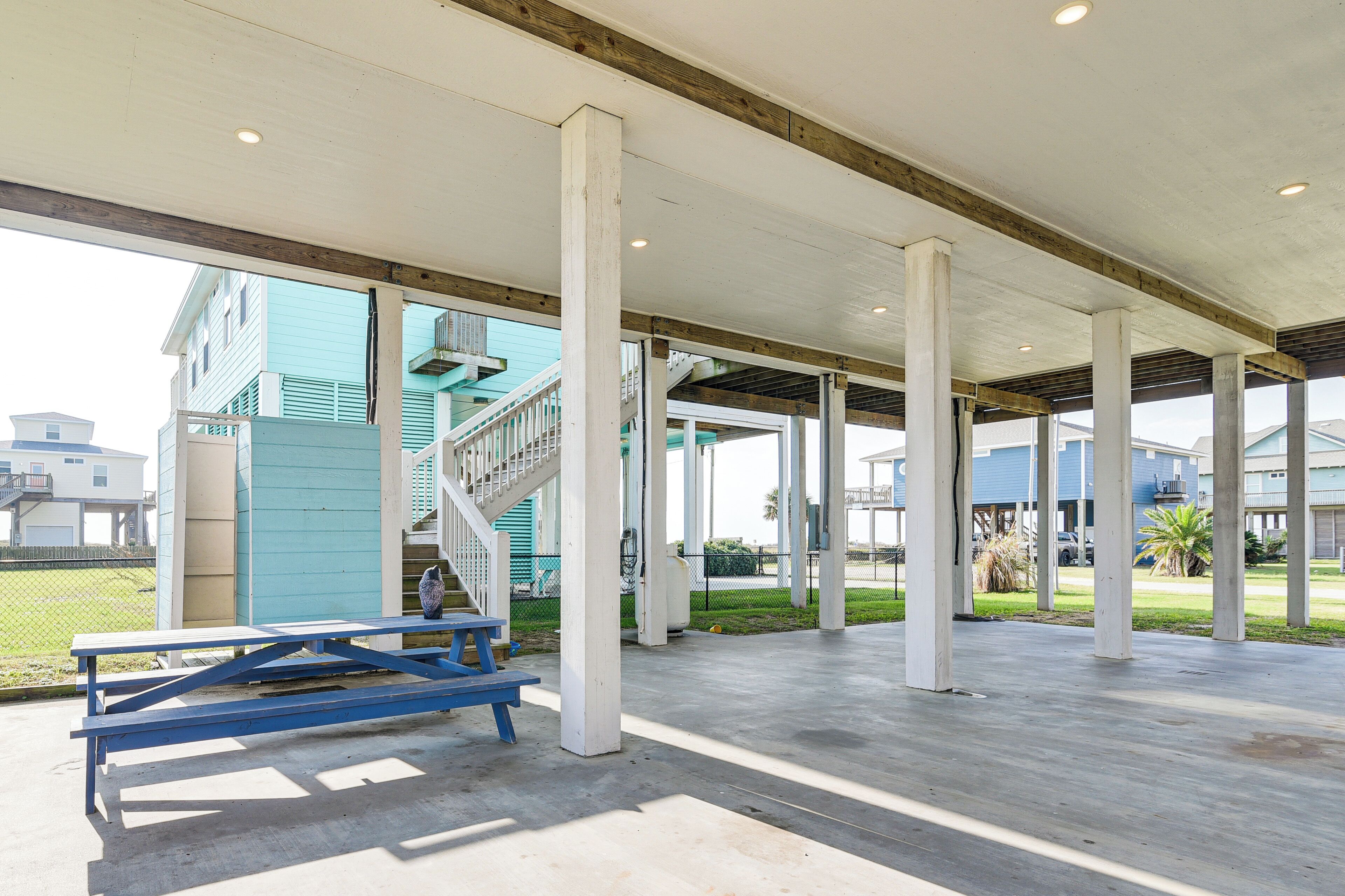 Un pavillon ouvert doté d’un plancher en béton et de piliers blancs, d’une table de pique-nique bleue et d’un escalier menant à un bâtiment aux bardages de couleur turquoise.