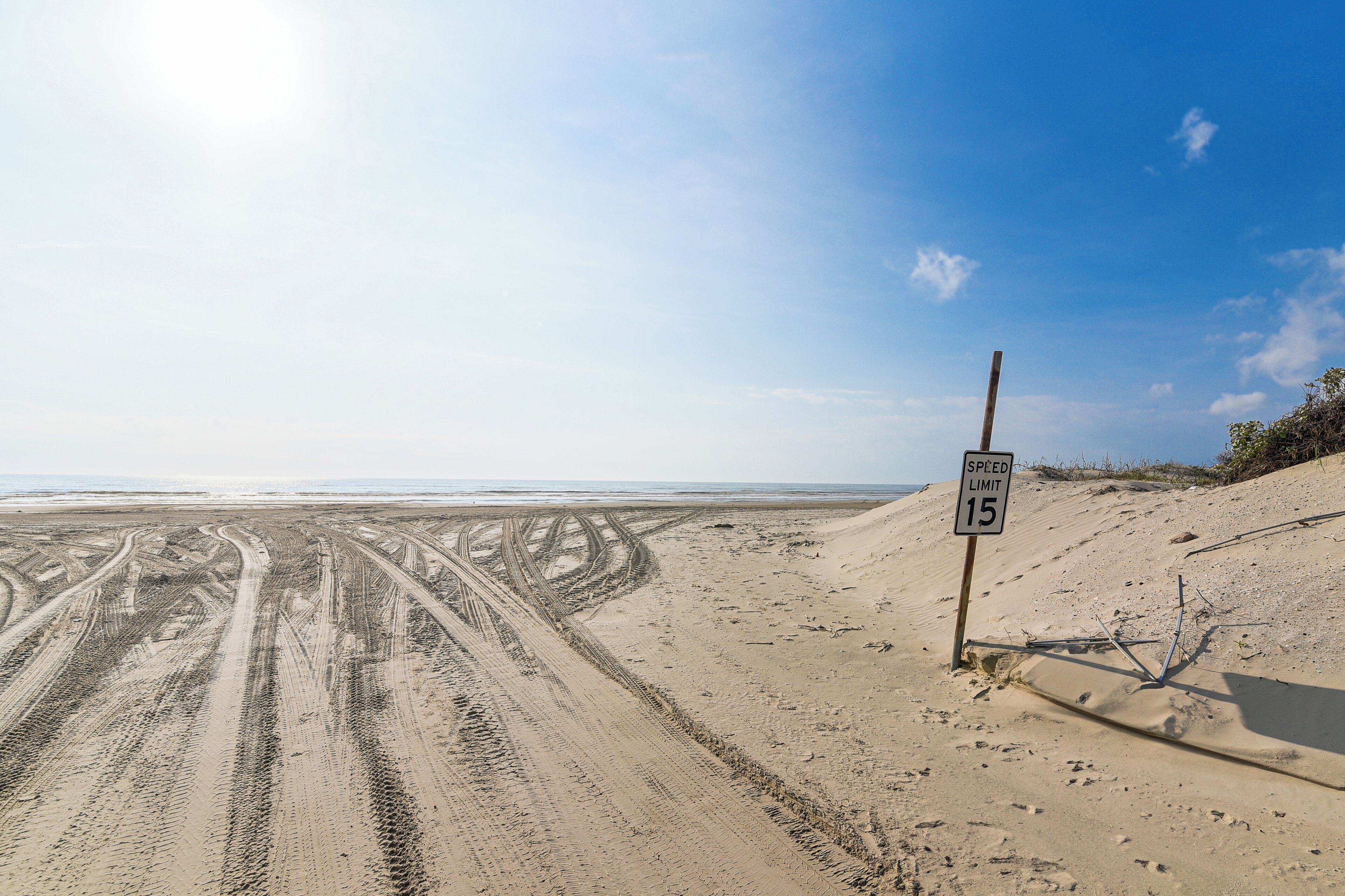 Praia arenosa com marcas de pneus e placa de velocidade indicando 24 quilômetros por hora.