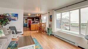 A spacious living room with a wooden floor, a dining area with chairs, a kitchen in the background, and large windows offering a view of the outside.