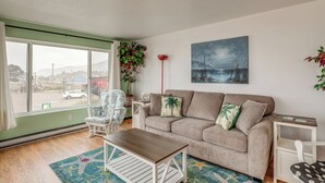 A living room with a sofa, a coffee table, and a chair. There is a window with a view of a street and mountains.