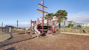 A playground with a slide and climbing structures, surrounded by a fence and a view of houses in the background.