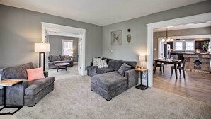 A living room with a grey sofa, a matching ottoman, and a coffee table. There is a dining area with a table and chairs. The room has a carpeted floor and a kitchen in the background. - Lakeside Garden Home - 1 Mile to Main Street! (Fort Wayne)