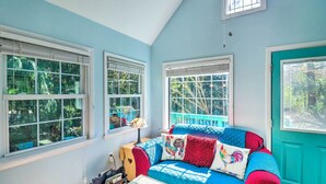 Una habitación con una pared azul, una puerta verde azulada y una cama con cubrecama azul y rojo acolchado. Hay tres ventanas con marcos y persianas blancos, y una pequeña mesa con una lámpara.
