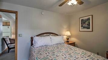 A bedroom with a bed, a nightstand with a lamp, a ceiling fan, a framed picture on the wall, and a chair in the corner.