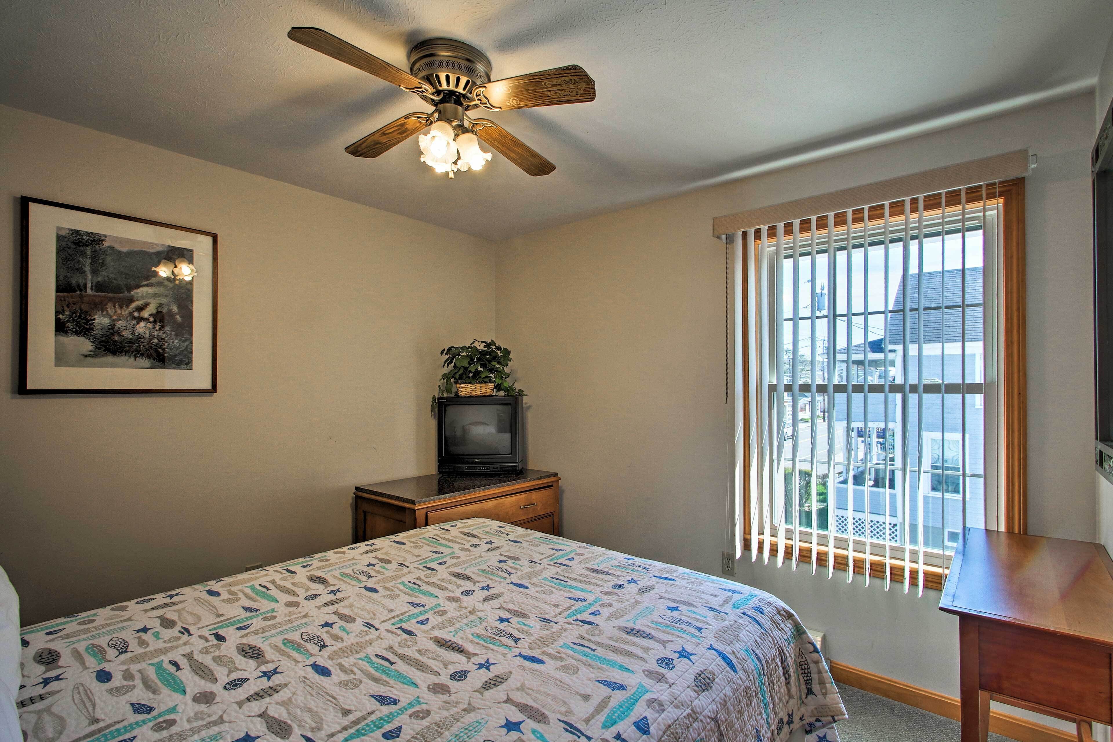 A bedroom with a bed, a ceiling fan, a small table with a plant, a TV on a stand, and a window with blinds.