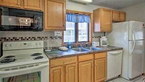 A kitchen with wooden cabinets, a white stove, a black microwave, a white refrigerator, and a window with a blue curtain. - Old Orchard Beach Apt - Walk to Beach & Pier! (Old Orchard Beach)